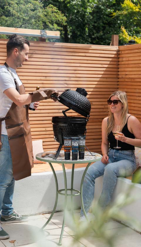 Man met BBQ schort naast kamado grill met sauzen in zonnige tuin
